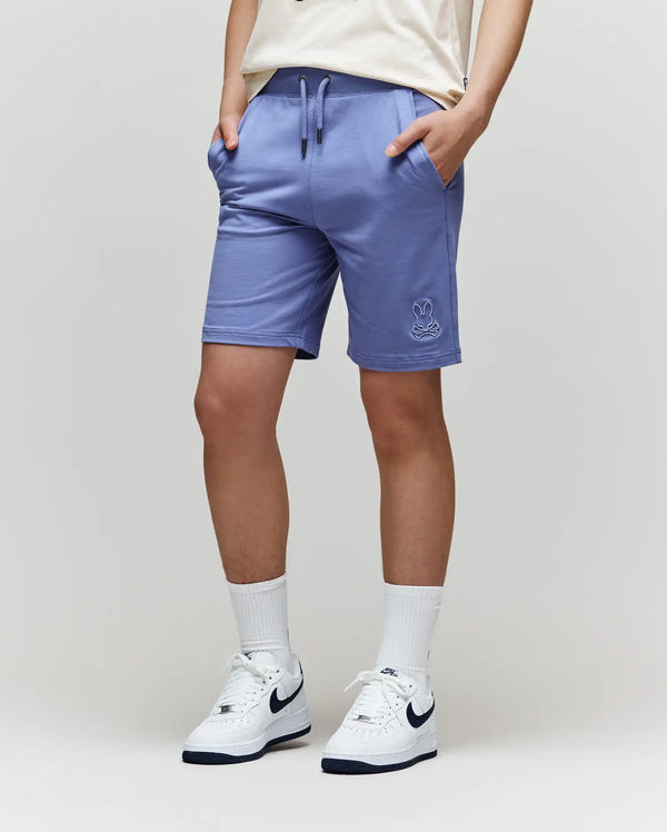 A person in light purple Psycho Bunny KIDS LEVANZO FRENCH TERRY SWEATSHORTS (B0R578F200), white crew socks, and white Nike sneakers with a dark blue swoosh stands against a plain light background, hands in pockets.