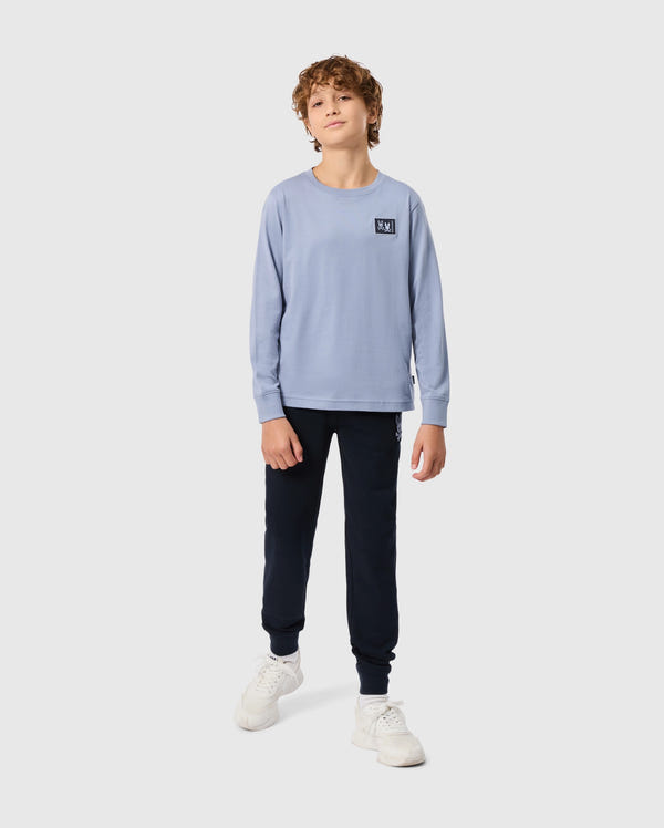 A young person with curly hair is in a Psycho Bunny KIDS MONROE LONG SLEEVE TEE (B0T975D200), light blue and pima cotton, featuring a small chest logo. Paired with dark joggers and white sneakers, the regular fit creates a cozy style against a plain white background.