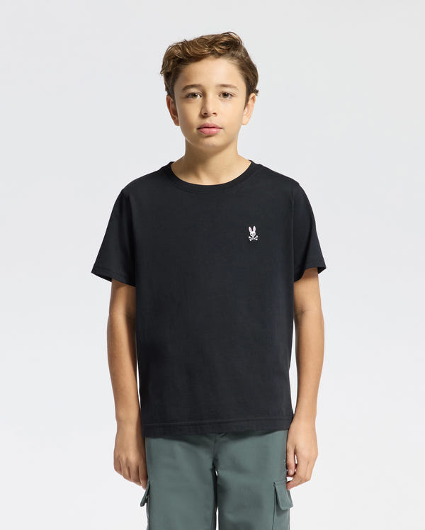 A young boy stands facing forward in a Psycho Bunny KIDS CLASSIC CREW NECK TEE - B0U014CRPC, made from 100% Pima cotton with a small white rabbit graphic on the chest. He pairs it with green cargo pants, ideal for back-to-school outfits against the plain white background.