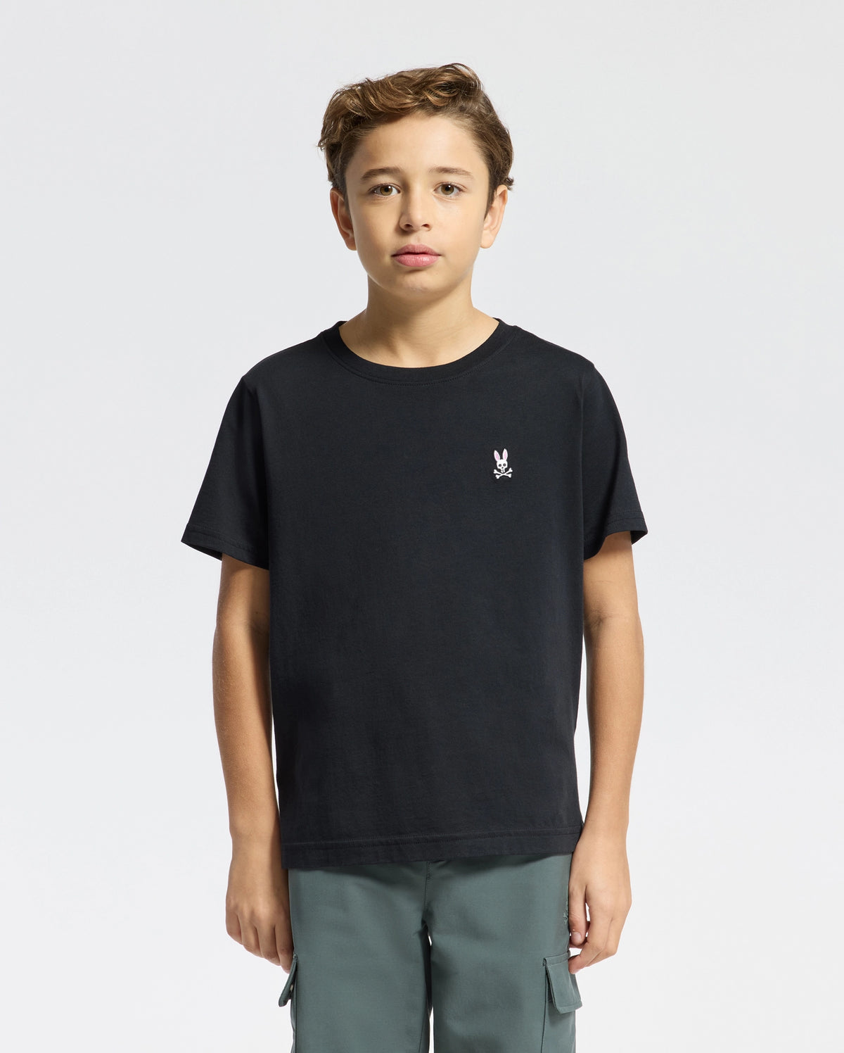 A young boy stands facing forward in a Psycho Bunny KIDS CLASSIC CREW NECK TEE - B0U014CRPC, made from 100% Pima cotton with a small white rabbit graphic on the chest. He pairs it with green cargo pants, ideal for back-to-school outfits against the plain white background.