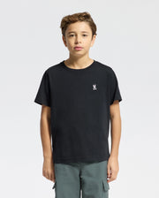 A young boy stands facing forward in a Psycho Bunny KIDS CLASSIC CREW NECK TEE - B0U014CRPC, made from 100% Pima cotton with a small white rabbit graphic on the chest. He pairs it with green cargo pants, ideal for back-to-school outfits against the plain white background.