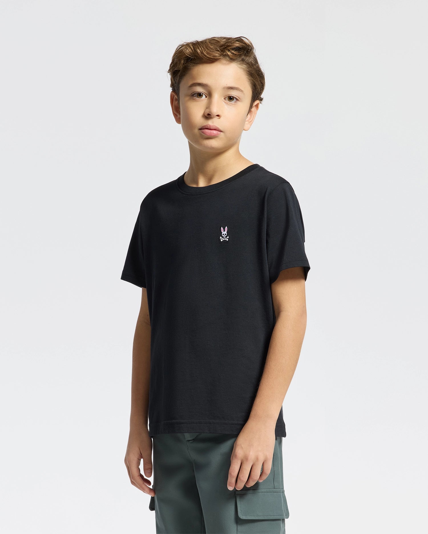 A young person with wavy hair stands against a plain background, wearing Psycho Bunny's black Kids Classic Crew Neck Tee (B0U014CRPC) featuring a small embroidered rabbit logo. Paired with green cargo pants, this short-sleeve tee offers comfort and style, perfect for back-to-school outfits.
