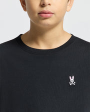Close-up of a person wearing Psycho Bunny's KIDS CLASSIC CREW NECK TEE - B0U014CRPC in black, featuring an embroidered pink rabbit with crossed bones. Made from 100% Pima cotton, it's perfect for back-to-school outfits. The shot shows them from neck down against a neutral background.