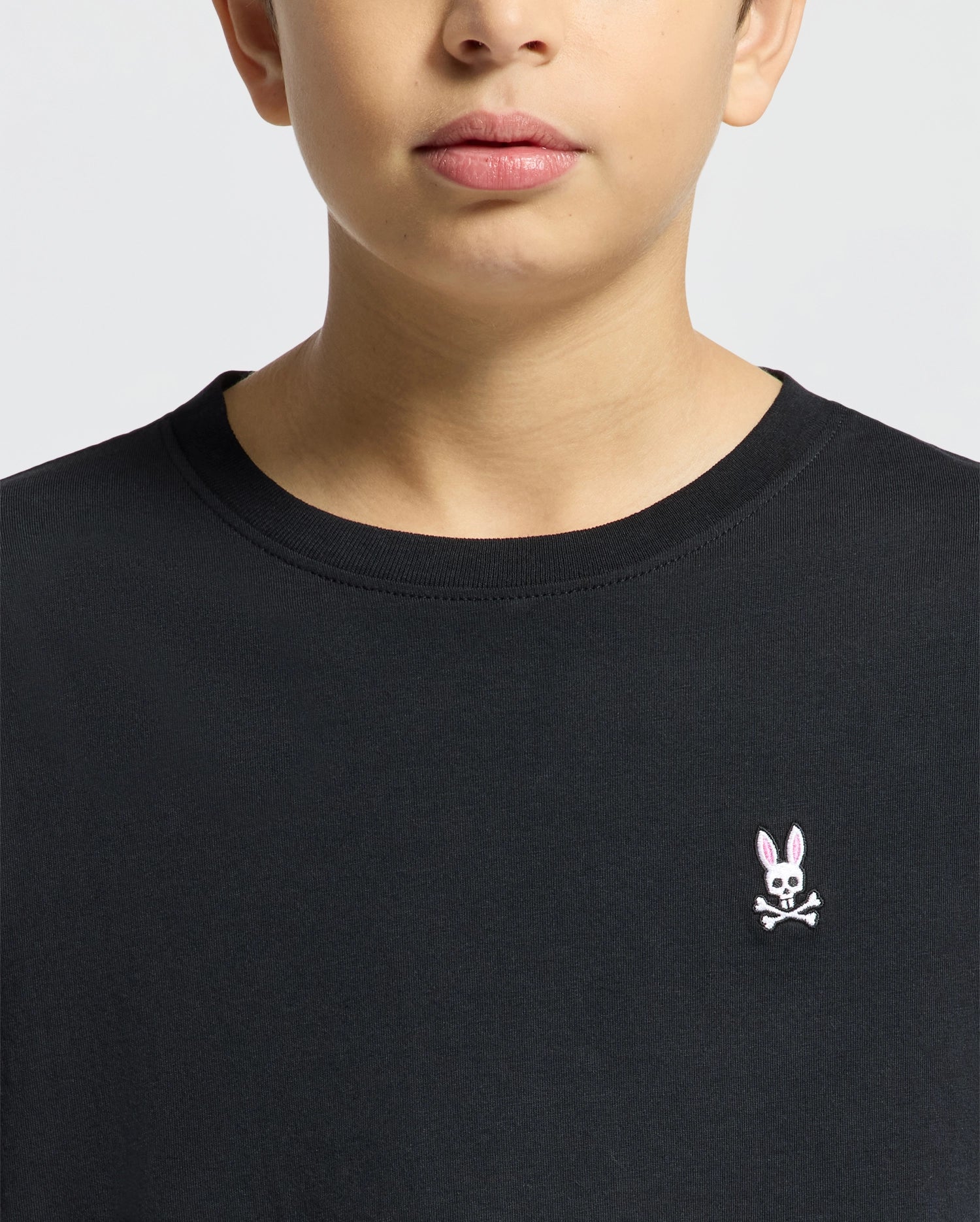 Close-up of a person wearing Psycho Bunny's KIDS CLASSIC CREW NECK TEE - B0U014CRPC in black, featuring an embroidered pink rabbit with crossed bones. Made from 100% Pima cotton, it's perfect for back-to-school outfits. The shot shows them from neck down against a neutral background.
