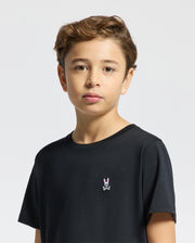 A boy with short, wavy brown hair wears a Psycho Bunny Kids Classic Crew Neck Tee (B0U014CRPC), made of 100% Pima cotton. It features a rabbit and crossbones logo and is perfect for back-to-school outfits. He stands against a plain white background, looking directly at the camera.