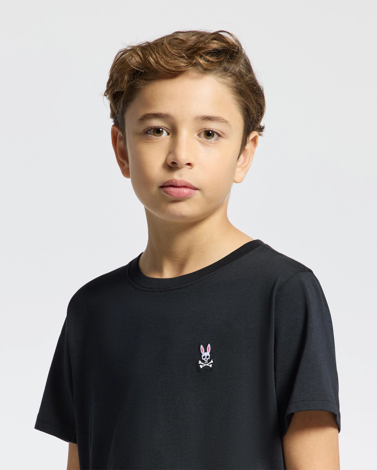 A boy with short, wavy brown hair wears a Psycho Bunny Kids Classic Crew Neck Tee (B0U014CRPC), made of 100% Pima cotton. It features a rabbit and crossbones logo and is perfect for back-to-school outfits. He stands against a plain white background, looking directly at the camera.