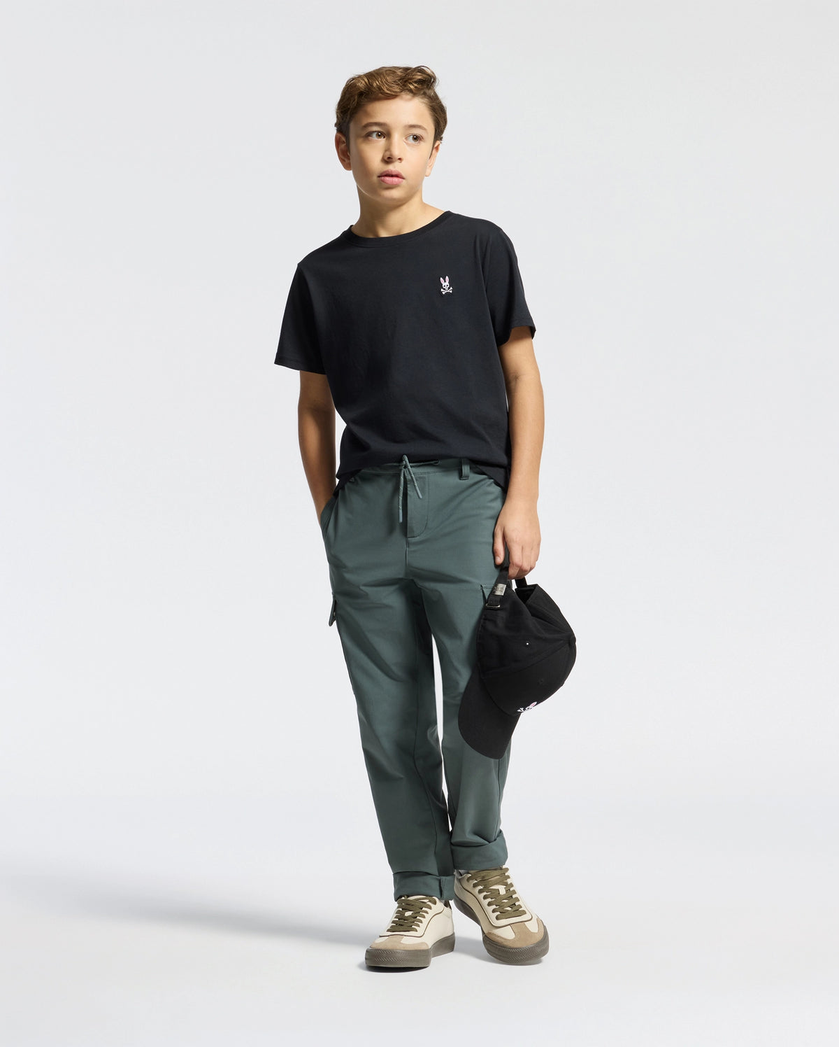 A young boy is wearing a Psycho Bunny KIDS CLASSIC CREW NECK TEE - B0U014CRPC made of 100% Pima cotton and green pants, holding a black cap in his left hand. He stands against a white background, looking to his left with white sneakers featuring beige and green accents—a perfect back-to-school look.