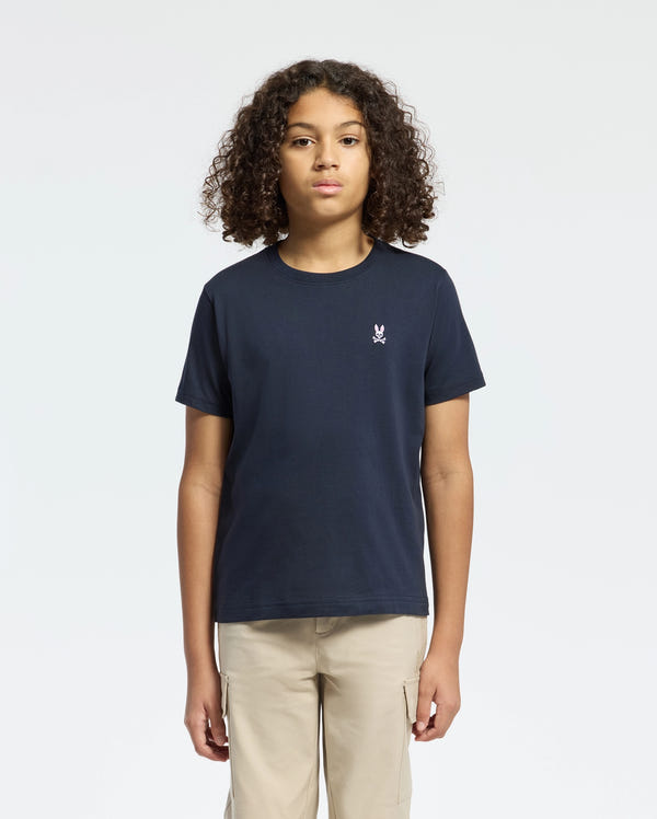A person with curly hair wears a Psycho Bunny KIDS CLASSIC CREW NECK TEE in navy blue, made from 100% Pima cotton with a small white logo on the chest. They're styled with beige pants against a plain white background—a timeless back-to-school look.