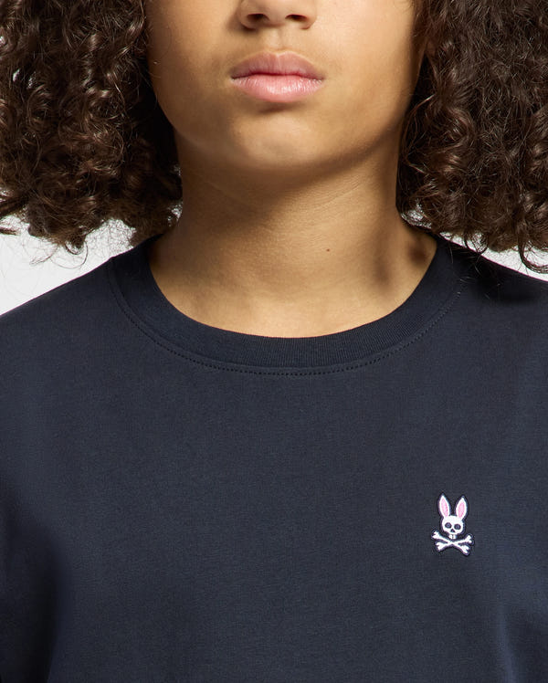 Someone with curly hair is wearing a dark KIDS CLASSIC CREW NECK TEE - B0U014CRPC by Psycho Bunny, featuring a small emblem of a pink-eared bunny and crossbones on the chest. Their partially visible face adds to the playful, stylish back-to-school vibe.