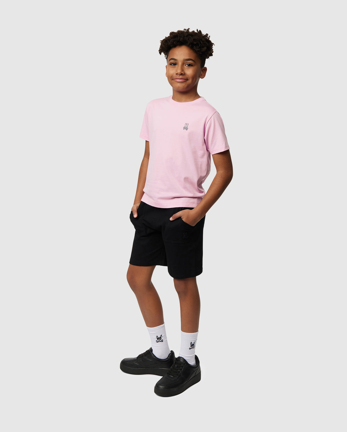 A young person stands confidently, wearing the Psycho Bunny KIDS CLASSIC CREW NECK TEE in pink, crafted from 100% Pima cotton. They pair it with black shorts and sneakers, casually resting their hands in their pockets to create a perfect back-to-school look against a plain gray background.