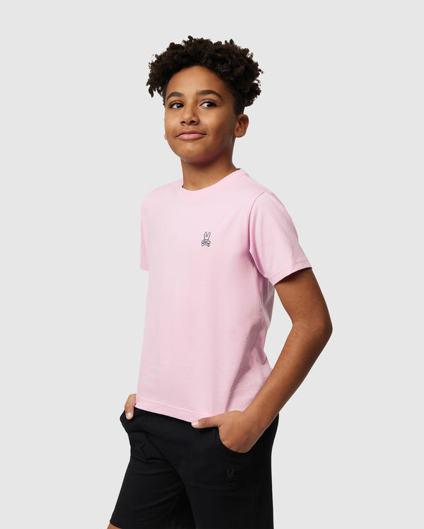A young individual wears a Psycho Bunny KIDS CLASSIC CREW NECK TEE in light pink, crafted from 100% Pima cotton, paired with black shorts. They stand with their hands in their pockets, offering a subtle smile against a plain gray background—an ideal back-to-school ensemble.