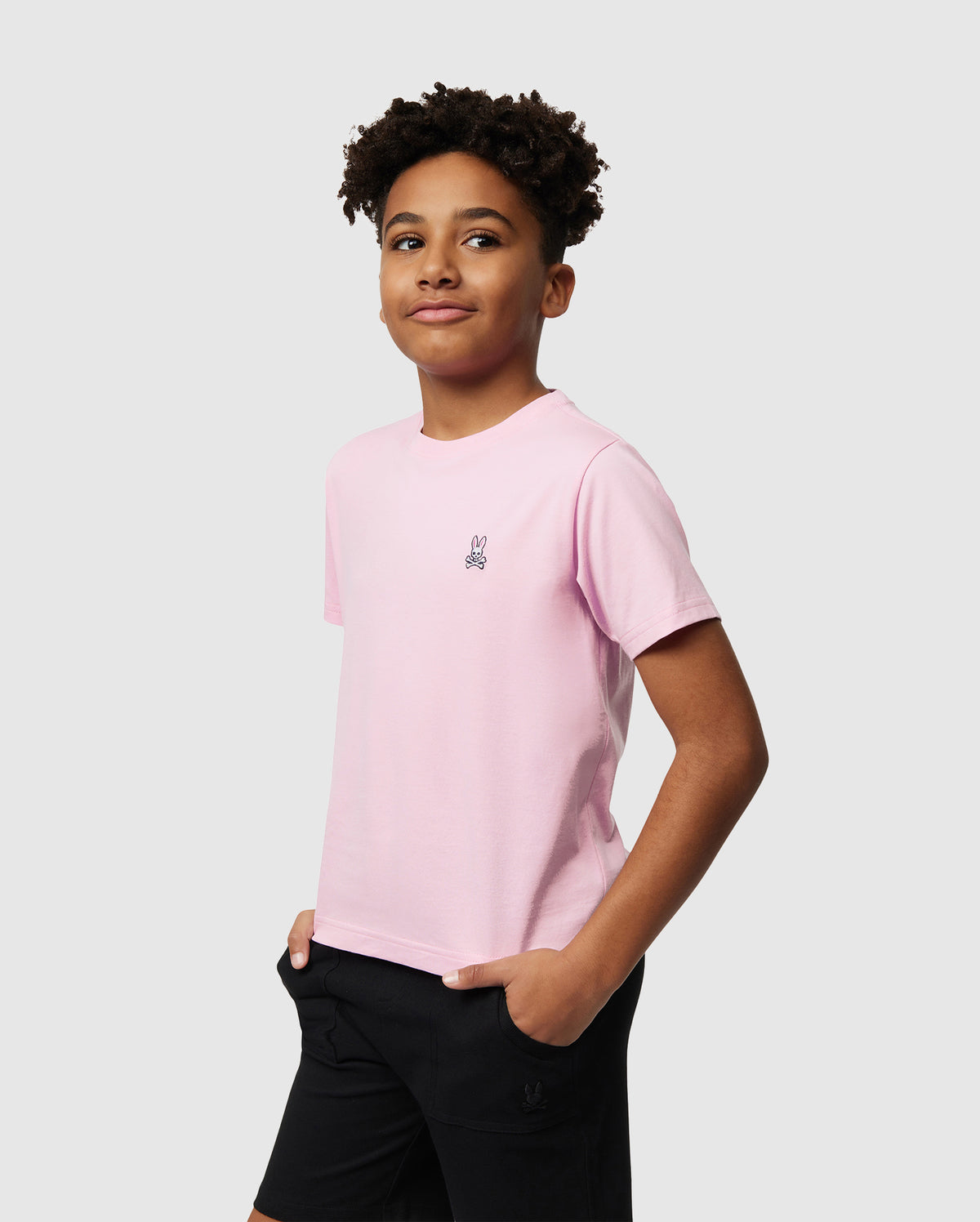A young individual wears a Psycho Bunny KIDS CLASSIC CREW NECK TEE in light pink, crafted from 100% Pima cotton, paired with black shorts. They stand with their hands in their pockets, offering a subtle smile against a plain gray background—an ideal back-to-school ensemble.