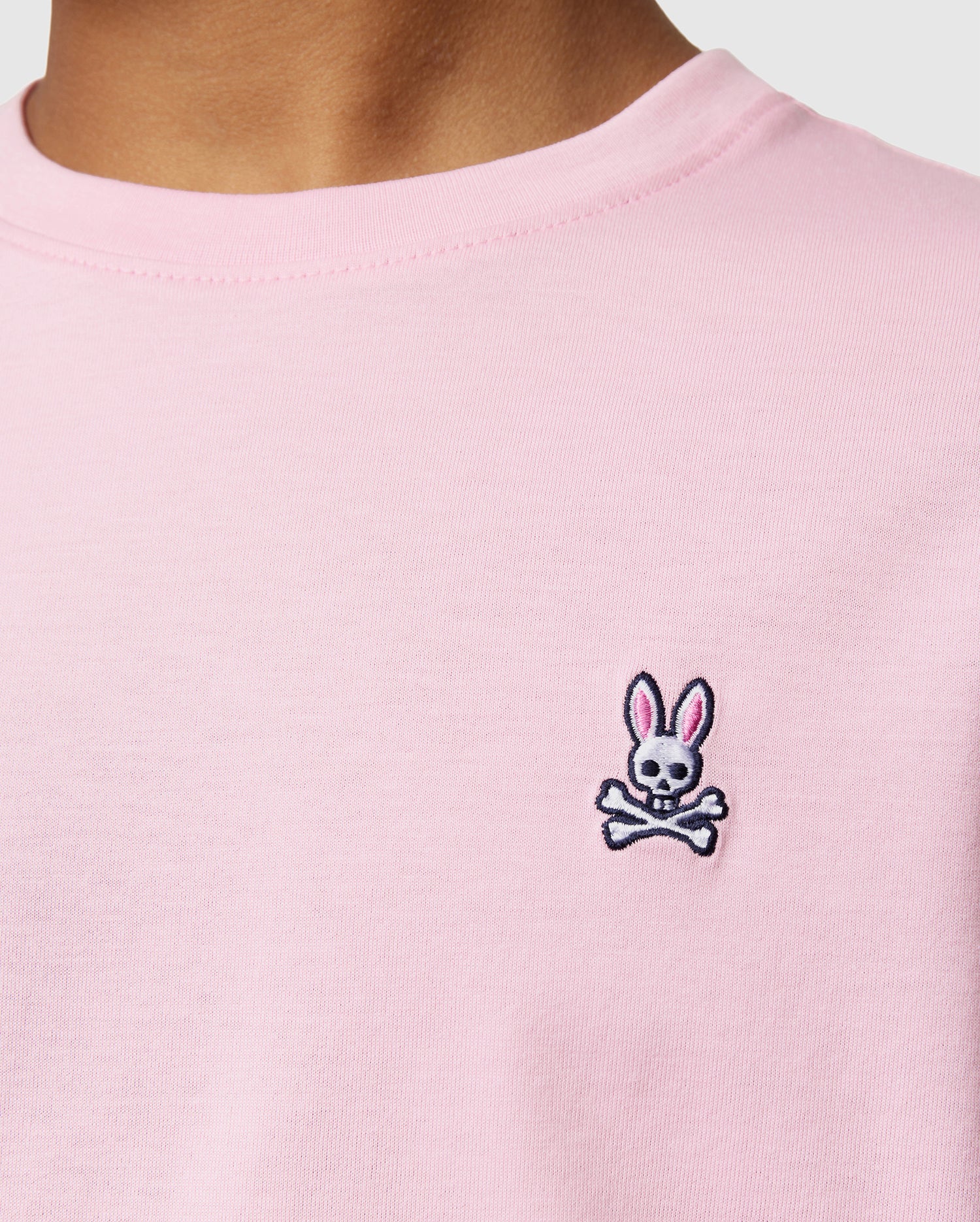 A person is wearing the KIDS CLASSIC CREW NECK TEE - B0U014CRPC by Psycho Bunny, a light pink shirt made from 100% Pima cotton with an embroidered rabbit skull and crossbones logo on the chest. This classic crew neck tee is ideal for back-to-school outfits and stands out beautifully against a plain white background.