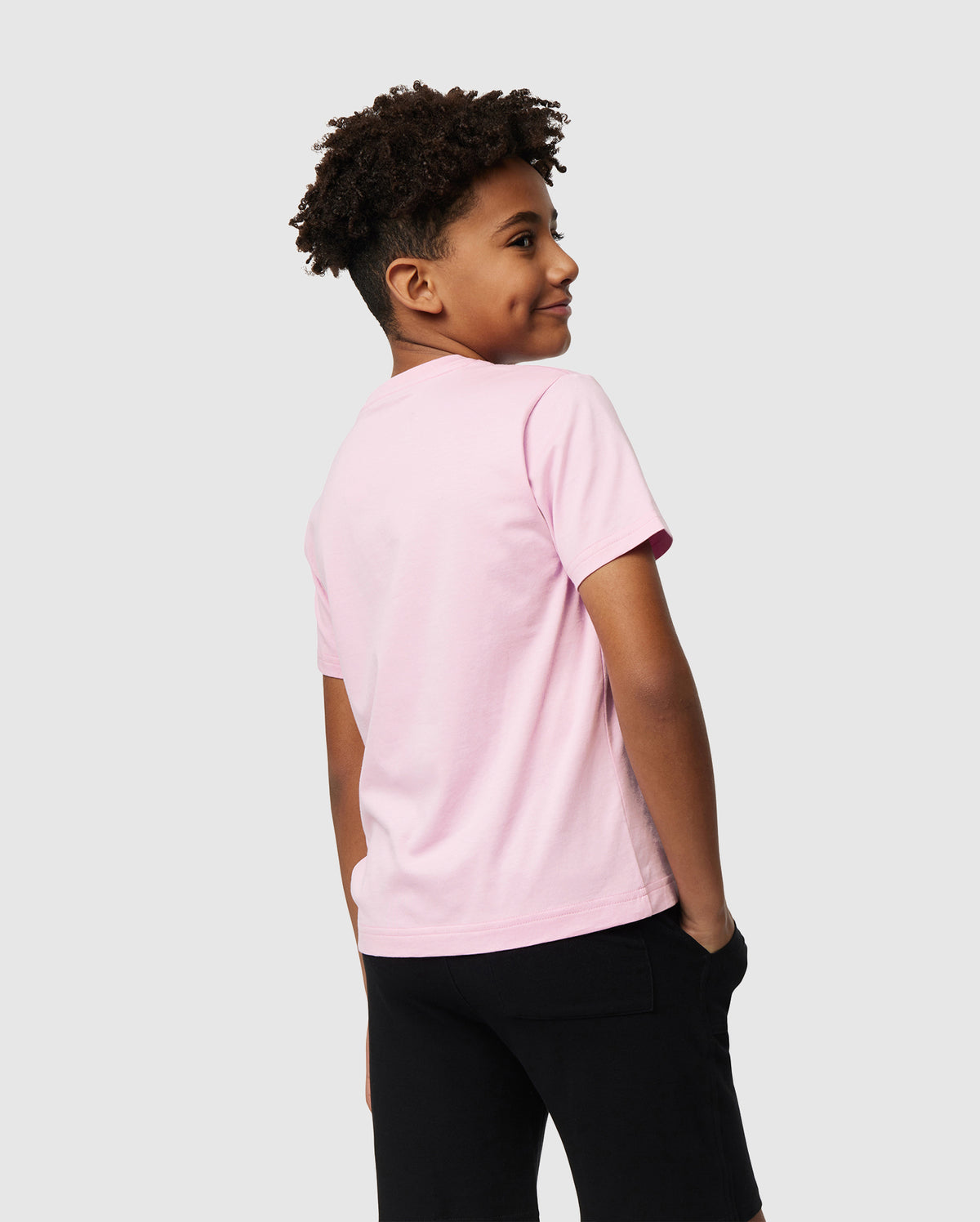 A young boy with curly hair stands smiling, looking over his shoulder. He is wearing a light pink Psycho Bunny Kids Classic Crew Neck Tee (B0U014CRPC) made from 100% Pima cotton and black shorts against a plain, light-colored background—perfect for back-to-school outfits.