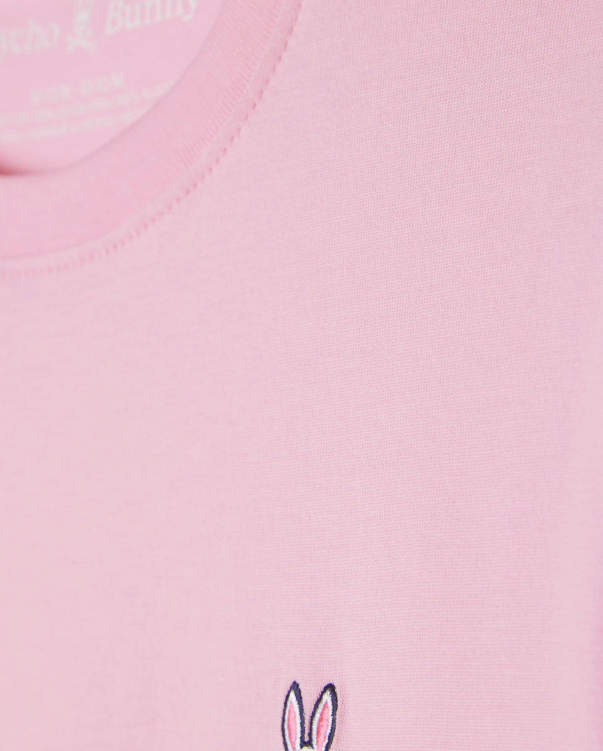 Close-up of the Kids Classic Crew Neck Tee (B0U014CRPC) by Psycho Bunny in pink, crafted from 100% Pima cotton. The fabric looks smooth and soft, and it features a small embroidered logo with bunny ears and facial details in black and white near the bottom edge—perfect for back-to-school outfits.