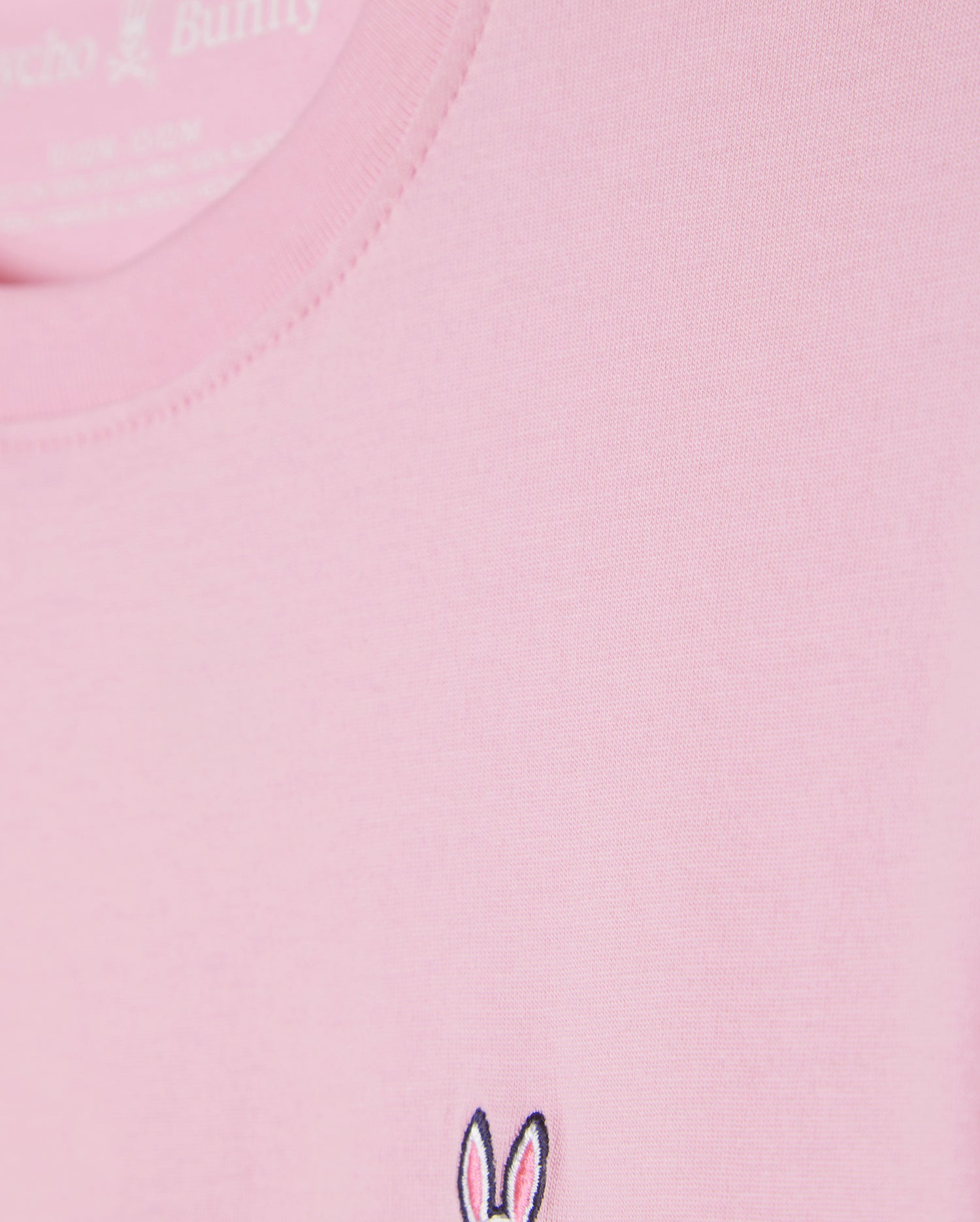 Close-up of the Kids Classic Crew Neck Tee (B0U014CRPC) by Psycho Bunny in pink, crafted from 100% Pima cotton. The fabric looks smooth and soft, and it features a small embroidered logo with bunny ears and facial details in black and white near the bottom edge—perfect for back-to-school outfits.