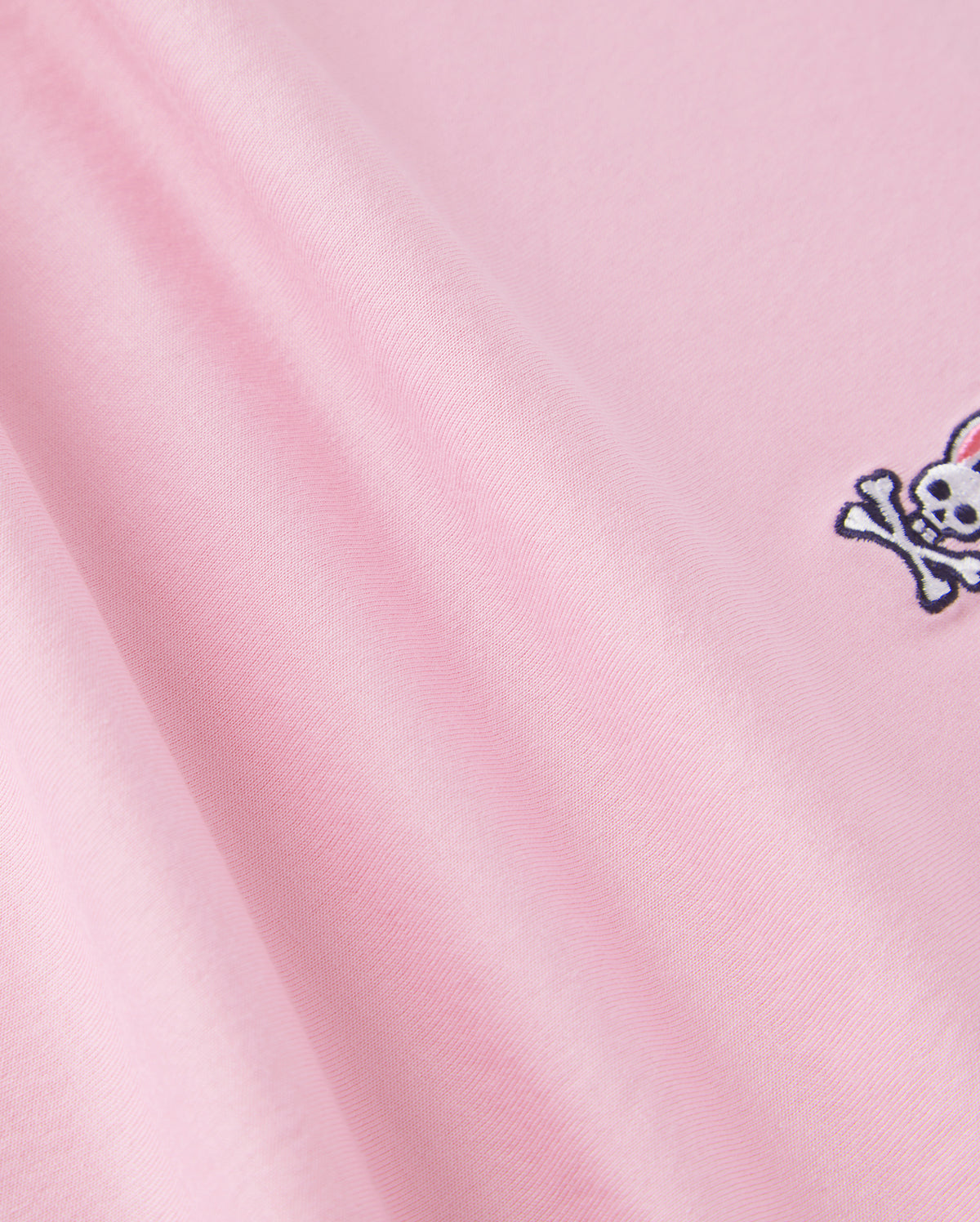 Close-up of pink fabric made from 100% Pima cotton, featuring a small embroidered design of a white skull and crossbones with bunny ears in the corner. This soft, smooth material with gentle folds is perfect for back-to-school outfits and ideal for Psycho Bunny's Kids Classic Crew Neck Tee - B0U014CRPC.