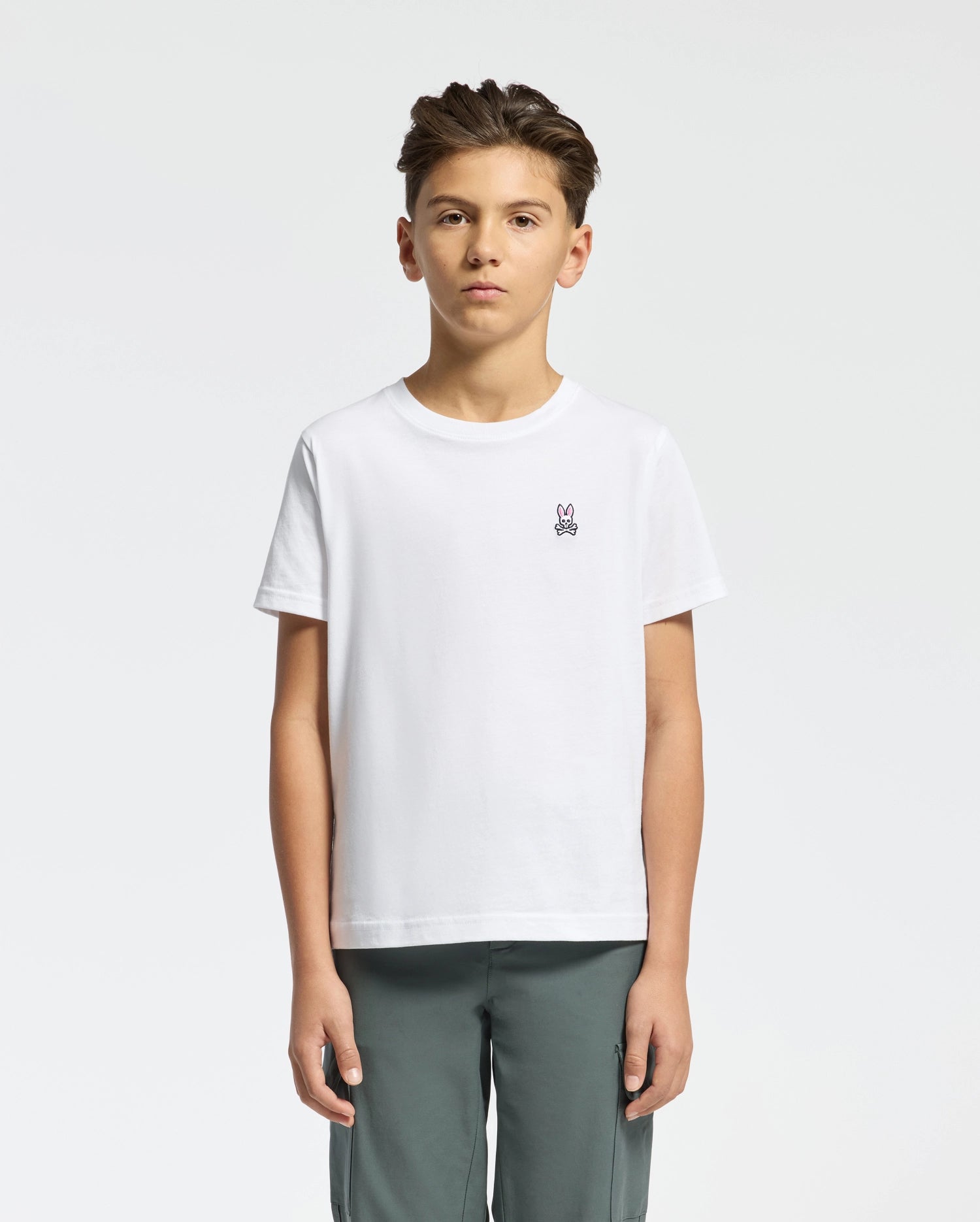 A young boy stands against a plain background, wearing Psycho Bunny's KIDS CLASSIC CREW NECK TEE - B0U014CRPC, made from 100% Pima cotton. The tee features a small bunny logo on the chest and is paired with gray pants. His neatly styled hair completes the back-to-school look.