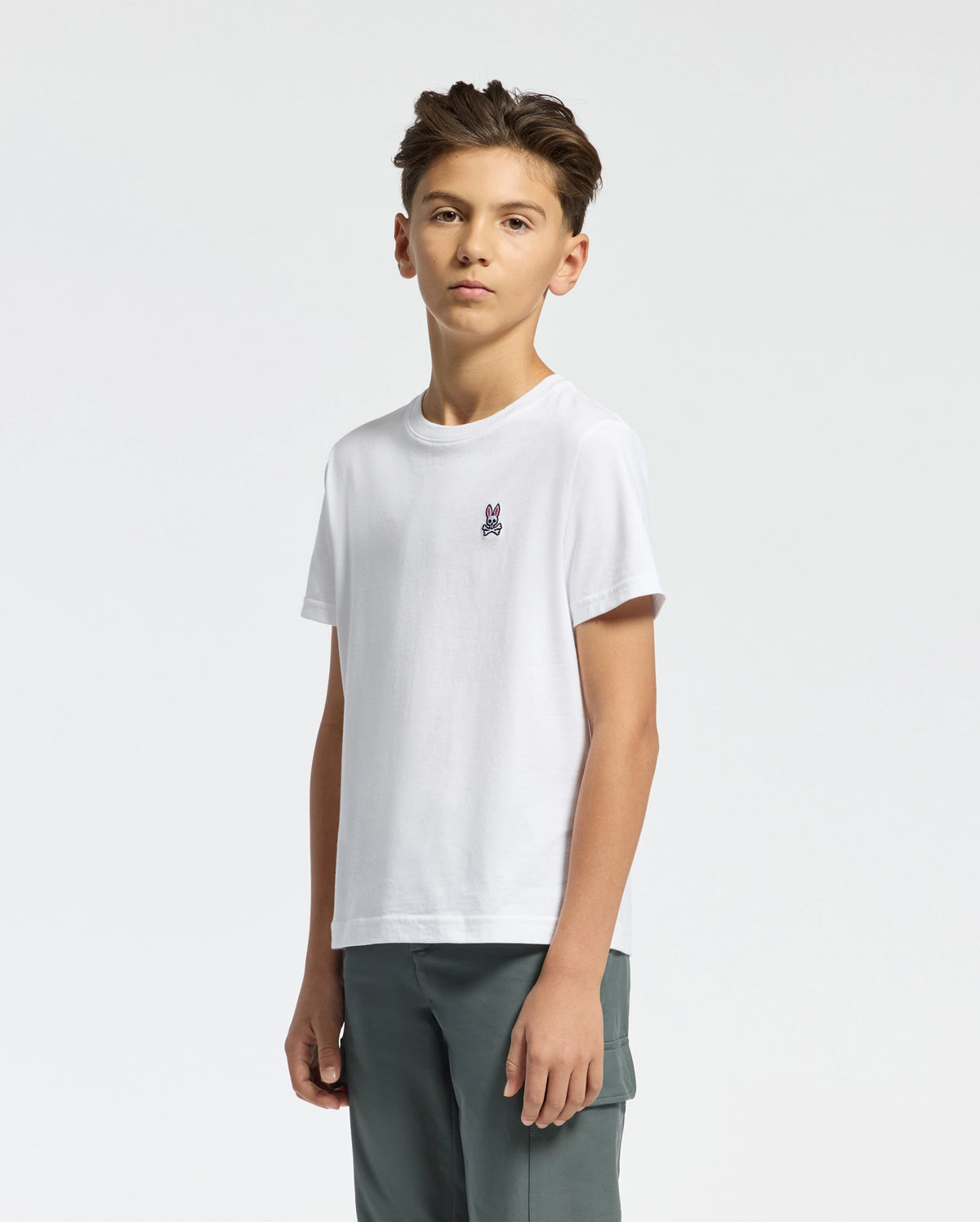 A young person with short brown hair wears Psycho Bunny's KIDS CLASSIC CREW NECK TEE - B0U014CRPC, crafted from 100% Pima cotton and featuring a chest graphic, paired with gray pants. Ideal for back-to-school outfits, they stand confidently against a plain white background.