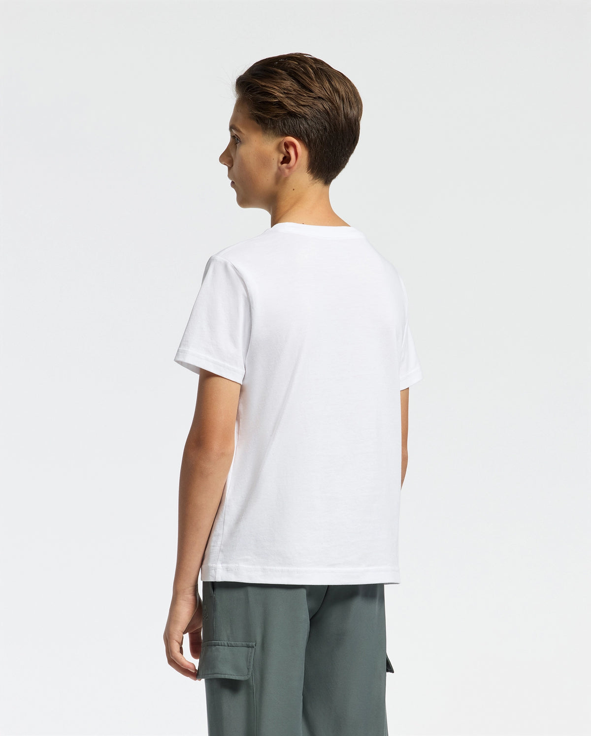 A boy with short brown hair stands sideways facing left, wearing the Psycho Bunny Kids Classic Crew Neck Tee (B0U014CRPC) in plain white and gray cargo pants against a white background—a perfect back-to-school outfit addition.