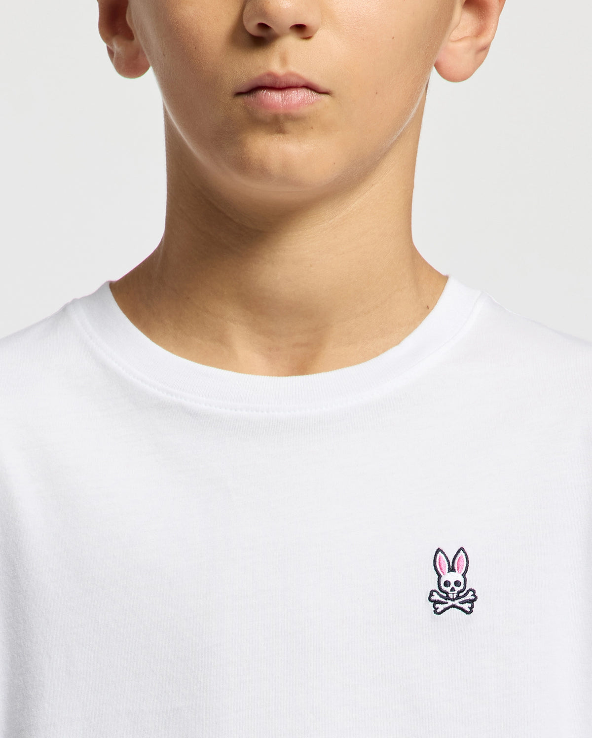 A person wears the KIDS CLASSIC CREW NECK TEE - B0U014CRPC by Psycho Bunny, featuring a small embroidered pink bunny and skull design on a plain white 100% Pima cotton T-shirt. It's perfect for adding unique flair to back-to-school outfits.