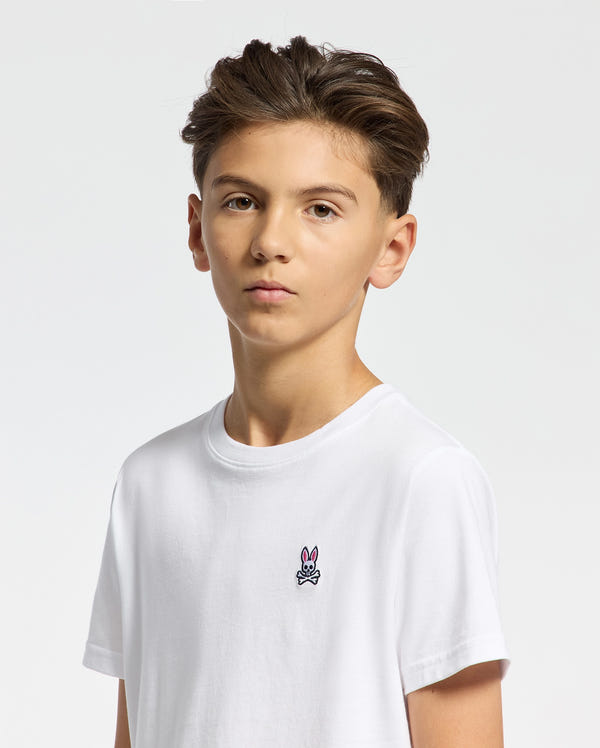 A young person with short brown hair wears the Psycho Bunny KIDS CLASSIC CREW NECK TEE - B0U014CRPC in crisp white, made from 100% Pima cotton, featuring a small bunny logo on the chest. Ideal for back-to-school outfits, its plain white background adds to its timeless appeal.