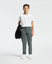A young person stands against a plain background, wearing the Psycho Bunny KIDS CLASSIC CREW NECK TEE in 100% Pima cotton, paired with green cargo pants. Holding a black jacket and sporting white sneakers, they exude a cool back-to-school vibe while looking directly at the camera.