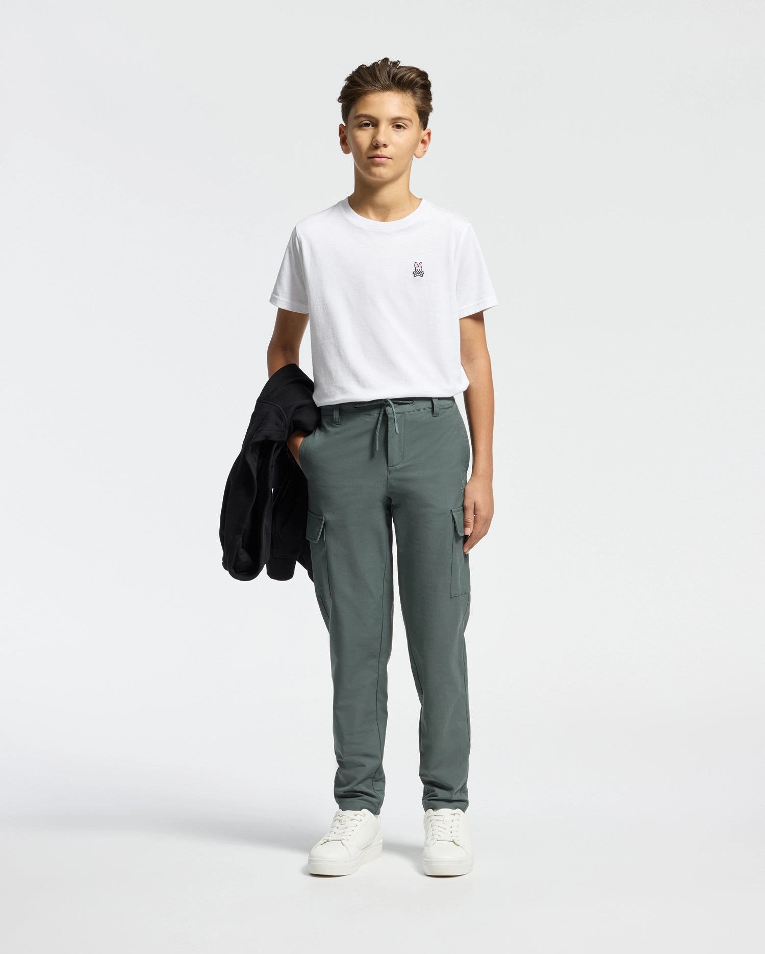 A young person stands against a plain background, wearing the Psycho Bunny KIDS CLASSIC CREW NECK TEE in 100% Pima cotton, paired with green cargo pants. Holding a black jacket and sporting white sneakers, they exude a cool back-to-school vibe while looking directly at the camera.