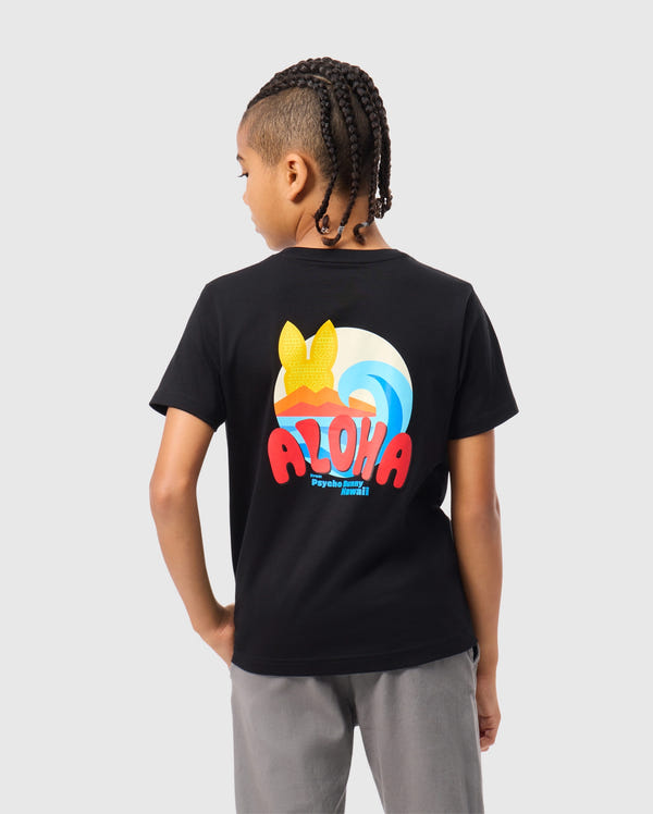 A child with braided hair wears the Psycho Bunny KIDS HAWAII CITY TEE - B0U169Y1PC, a Pima cotton shirt in black, showcasing vibrant mountains and waves with "Aloha" in bold letters. It stands out against a light gray backdrop, capturing the volcanic island spirit.