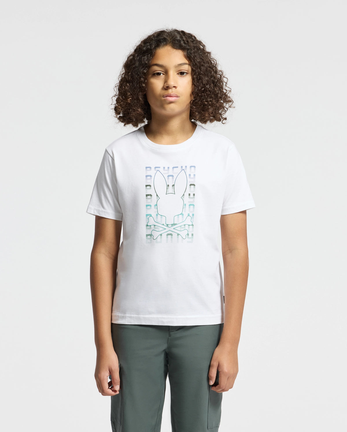 A person with curly hair is wearing the KIDS CORTES GRAPHIC TEE - B0U328E200 by Psycho Bunny, crafted from 100% cotton and showcasing a stylized rabbit graphic, stands against a plain background. They are also wearing dark pants.