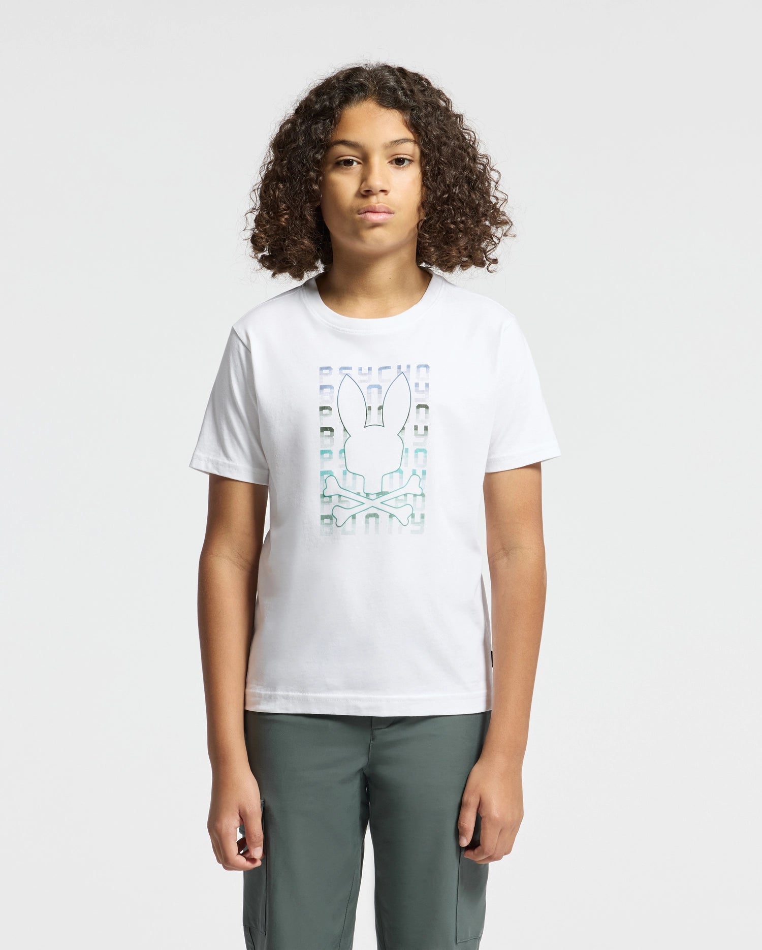 A person with curly hair is wearing the KIDS CORTES GRAPHIC TEE - B0U328E200 by Psycho Bunny, crafted from 100% cotton and showcasing a stylized rabbit graphic, stands against a plain background. They are also wearing dark pants.