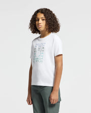 A person with curly hair is dressed in a stylish Psycho Bunny KIDS CORTES GRAPHIC TEE - B0U328E200, made from 100% cotton, paired with dark green pants, standing against a plain white background.