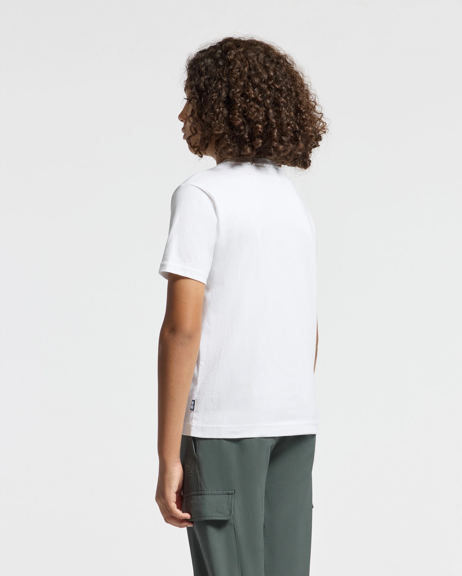 A person with curly hair, standing and facing away, is showcasing a fashionable KIDS CORTES GRAPHIC TEE - B0U328E200 by Psycho Bunny. They are wearing green pants, and the tee appears to be soft and comfortable, suggesting it might be made from 100% cotton. The background is plain and light-colored.