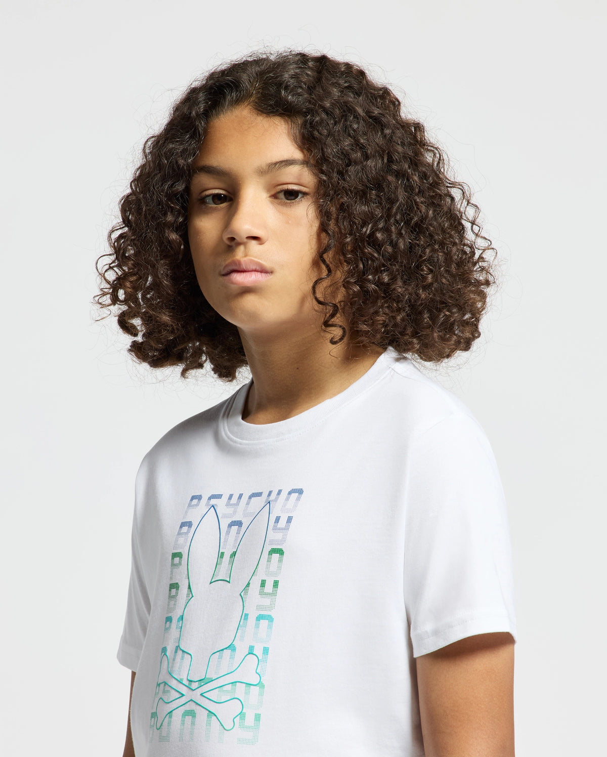 A child with curly hair is wearing a stylish Psycho Bunny KIDS CORTES GRAPHIC TEE - B0U328E200, made from 100% cotton. The tee features a rabbit and crossbones design on a white backdrop, set against a simple, light-colored background.