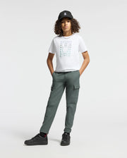 A person with curly hair is wearing a black cap, the stylish Psycho Bunny KIDS CORTES GRAPHIC TEE - B0U328E200 made from 100% cotton, and green cargo pants. They stand with hands in pockets, sporting black sneakers against a plain white background.