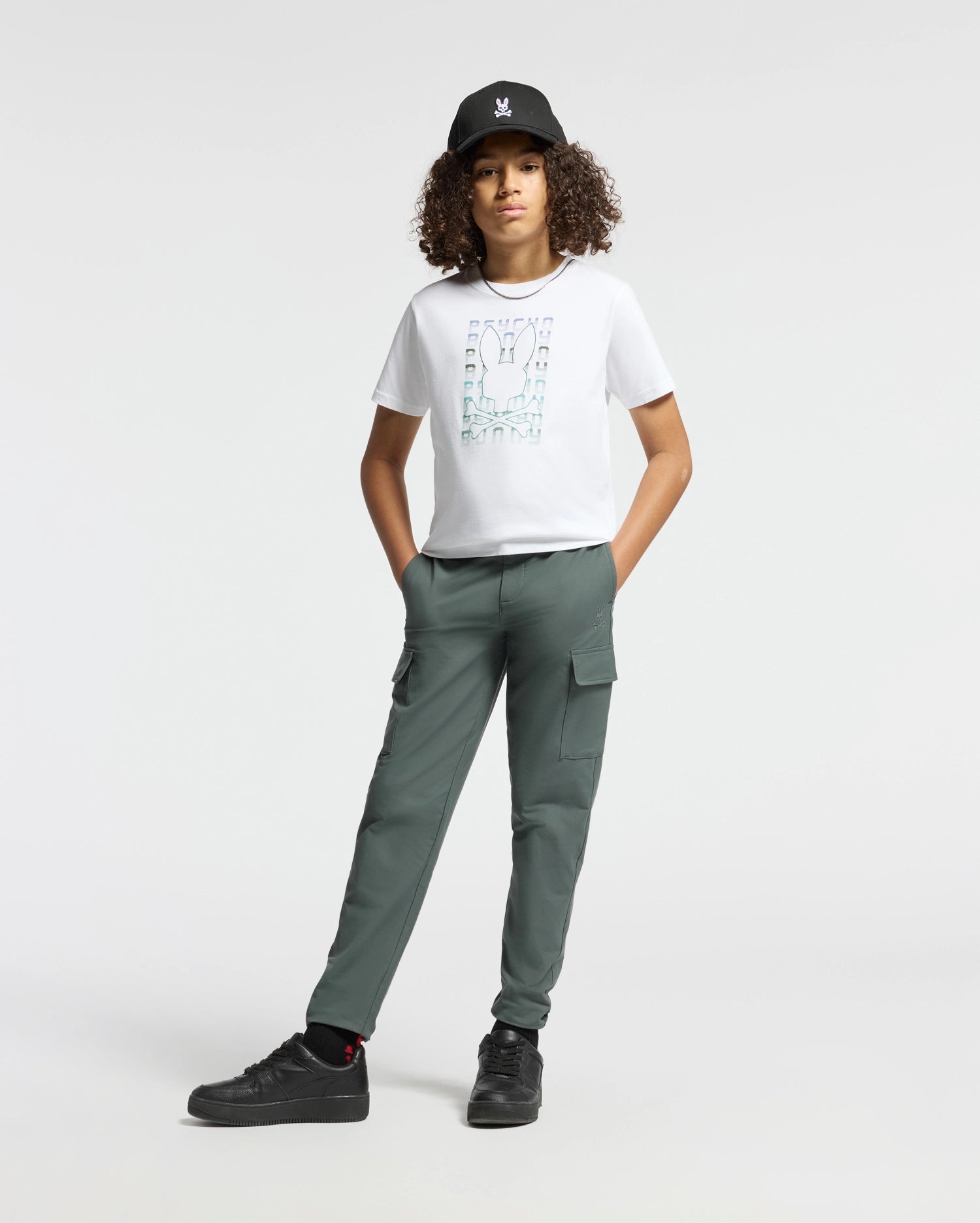 A person with curly hair is wearing a black cap, the stylish Psycho Bunny KIDS CORTES GRAPHIC TEE - B0U328E200 made from 100% cotton, and green cargo pants. They stand with hands in pockets, sporting black sneakers against a plain white background.