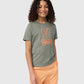 A person with curly hair smiles while wearing a green, regular fit KIDS FLOYD GRAPHIC TEE - B0U338B2TS by Psycho Bunny featuring an embroidered bunny outline and crossbones design and peach-colored shorts. The background is plain light grey.
