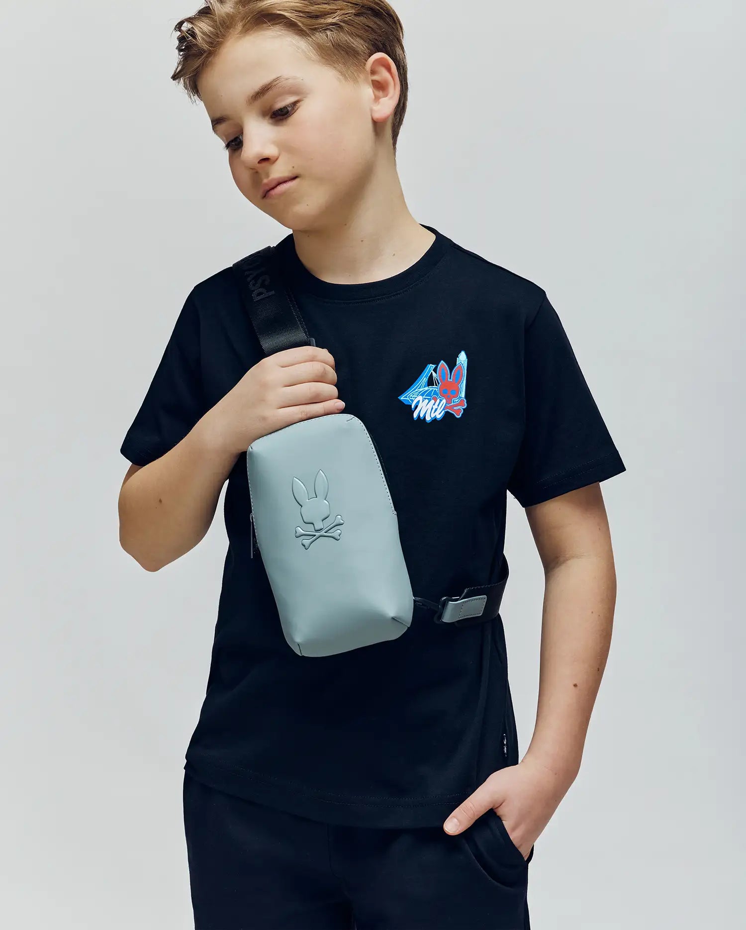 A boy wears the Psycho Bunny KIDS MONTREAL TEE (B0U381F200) with a colorful graphic, black shorts, and a light gray crossbody bag featuring the bunny logo. He stands against a plain background, looking down with a relaxed expression.