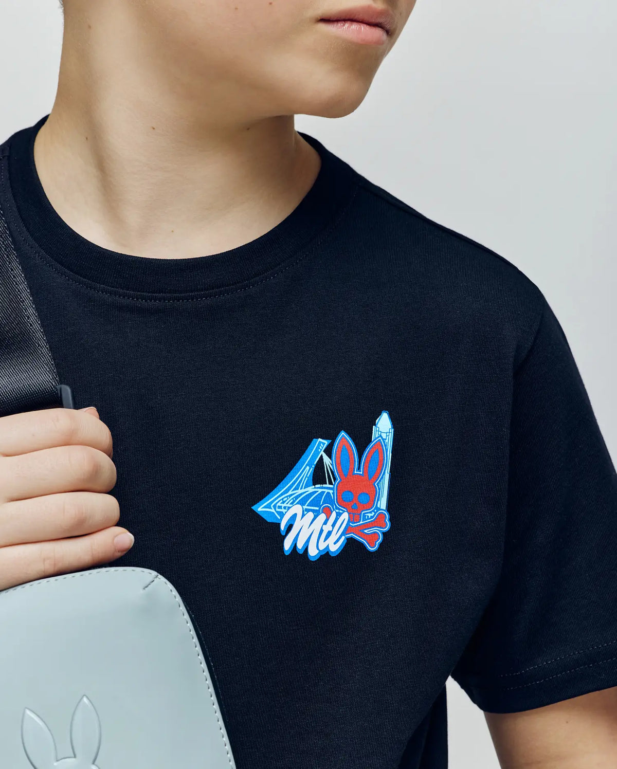 A young person wearing the Psycho Bunny KIDS MONTREAL TEE (B0U381F200)—a soft black Pima cotton shirt with a vibrant bunny and skull chest graphic—holds a light gray bag over their shoulder, looking to the side with a partially visible face.