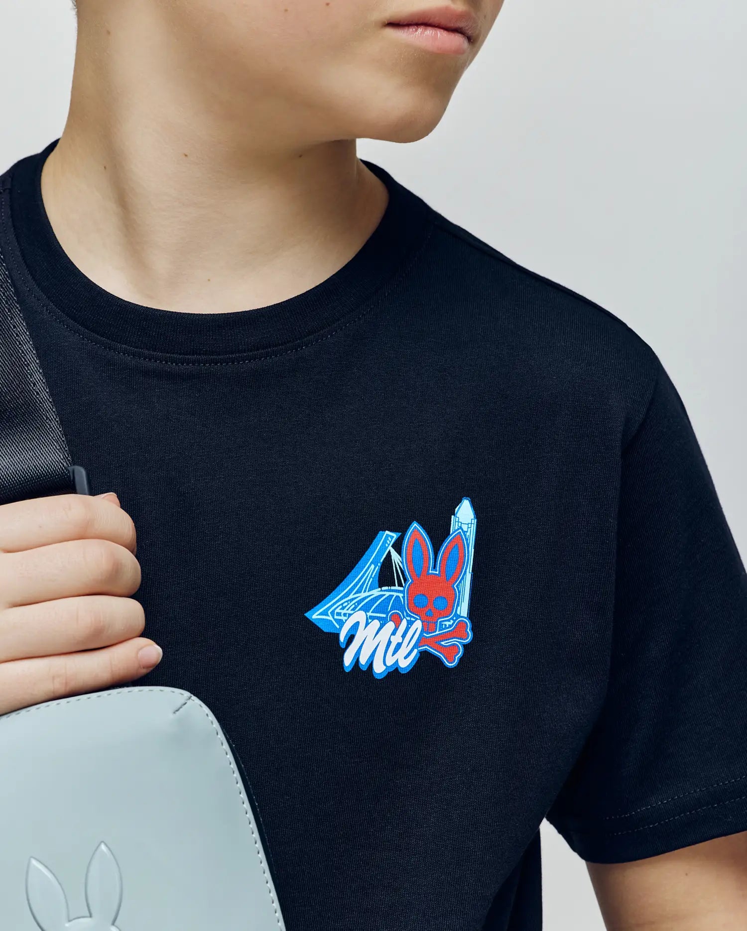 A young person wearing the Psycho Bunny KIDS MONTREAL TEE (B0U381F200)—a soft black Pima cotton shirt with a vibrant bunny and skull chest graphic—holds a light gray bag over their shoulder, looking to the side with a partially visible face.