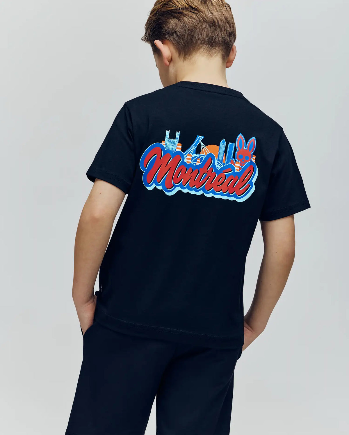 A young boy faces away from the camera, wearing the Psycho Bunny KIDS MONTREAL TEE (B0U381F200), which showcases a vibrant Montreal skyline graphic and bold “Montreal” lettering on the back.