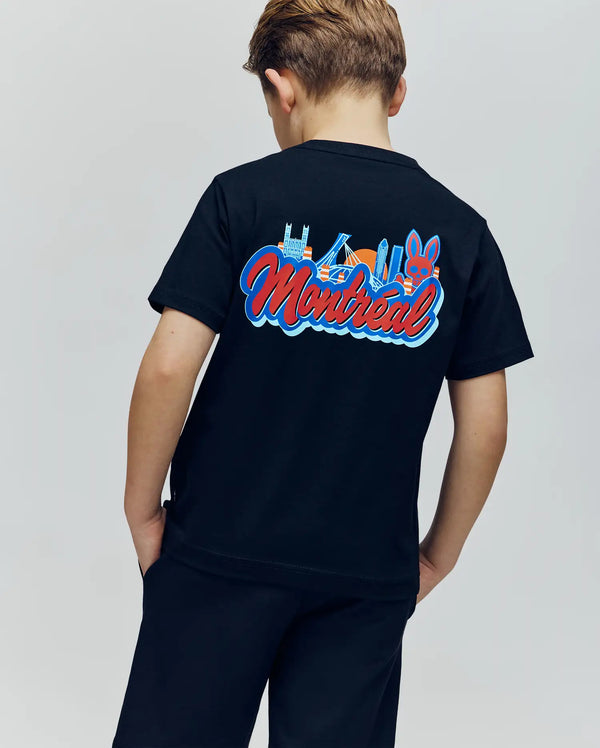 A young boy faces away from the camera, wearing the Psycho Bunny KIDS MONTREAL TEE (B0U381F200), which showcases a vibrant Montreal skyline graphic and bold “Montreal” lettering on the back.