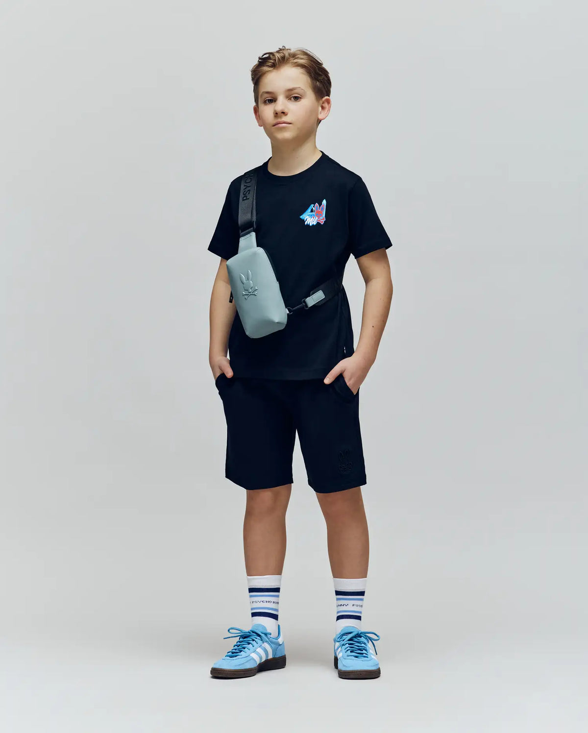 A boy stands against a plain background, wearing the Psycho Bunny KIDS MONTREAL TEE (B0U381F200) in black with shorts, blue and white sneakers, striped socks, and a light gray crossbody bag. He looks forward neutrally, hands in his pockets.