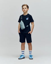A boy stands against a plain background, wearing the Psycho Bunny KIDS MONTREAL TEE (B0U381F200) in black with shorts, blue and white sneakers, striped socks, and a light gray crossbody bag. He looks forward neutrally, hands in his pockets.