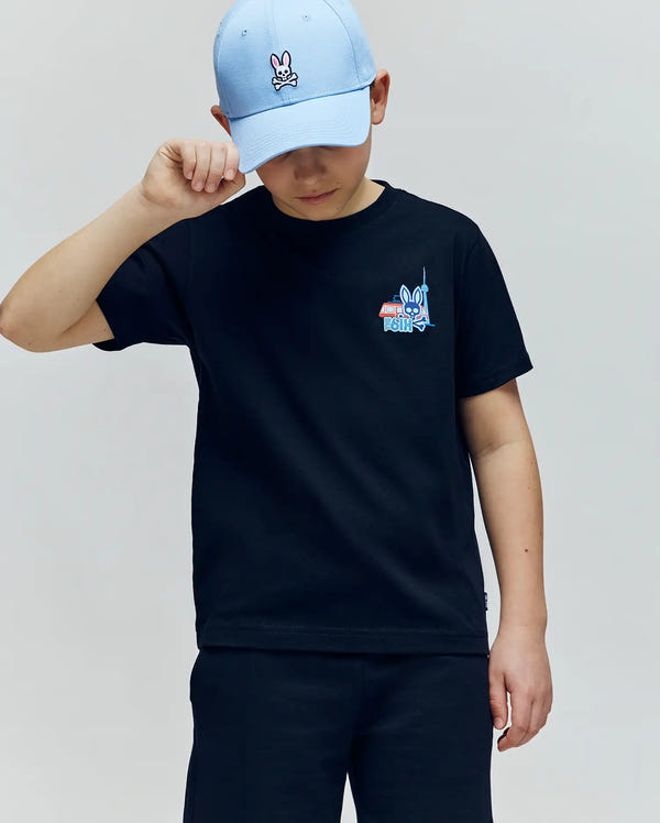 A child in a light blue cap and a Psycho Bunny KIDS TORONTO TEE (B0U383F200), made from soft Pima cotton with playful graphics, stands against a plain background, looking down and touching the brim of the cap.