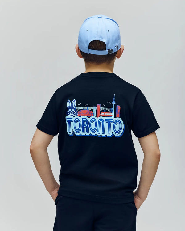 A child in a light blue cap wears the Psycho Bunny KIDS TORONTO TEE (B0U383F200), made from soft Pima cotton and featuring vibrant prints of a bunny, city buildings, and the CN Tower, standing with their back to the camera.