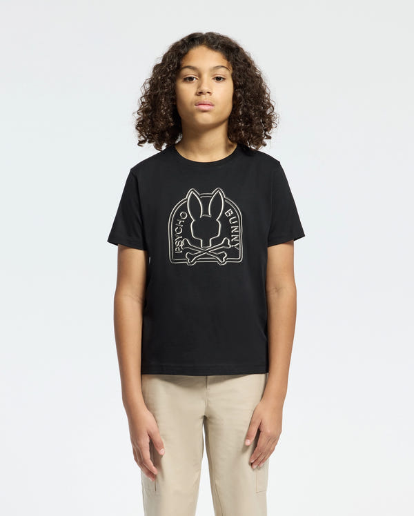 A person with curly hair is wearing a black Kids Melbourne Graphic Tee (B0U410F200) made of Pima cotton by Psycho Bunny, featuring a white bunny graphic above crossed golf clubs and "Psycho Bunny" text. They are also in beige pants, standing against a plain white background.
