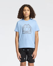 A person with curly hair wears a Psycho Bunny Kids Melbourne Graphic Tee (B0U410F200) in light blue, crafted from eco-friendly Peruvian Pima cotton, with a bunny and crossed bones graphic. They pair it with black shorts featuring small white Psycho Bunny logos against a plain white background.