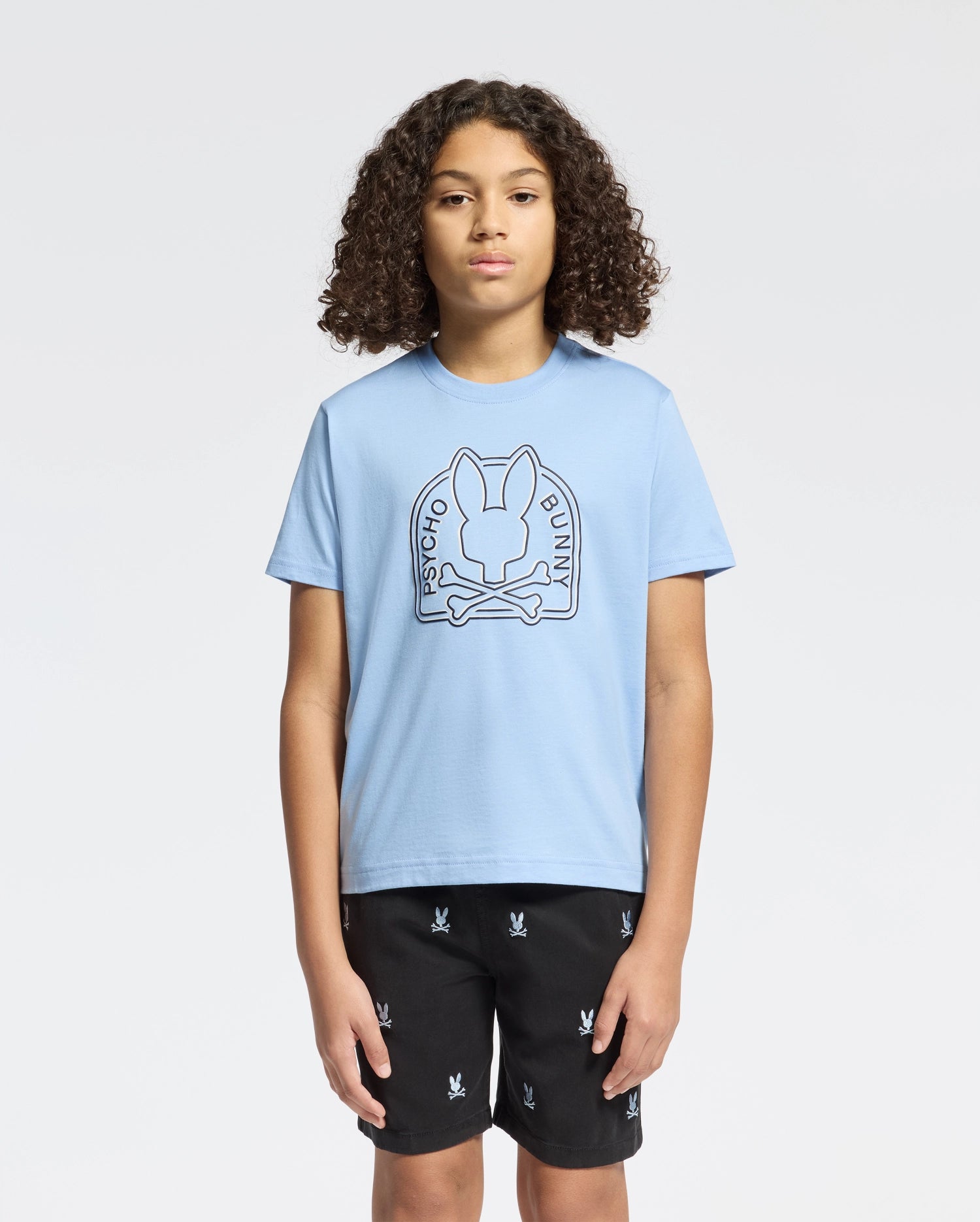 A person with curly hair wears a Psycho Bunny Kids Melbourne Graphic Tee (B0U410F200) in light blue, crafted from eco-friendly Peruvian Pima cotton, with a bunny and crossed bones graphic. They pair it with black shorts featuring small white Psycho Bunny logos against a plain white background.