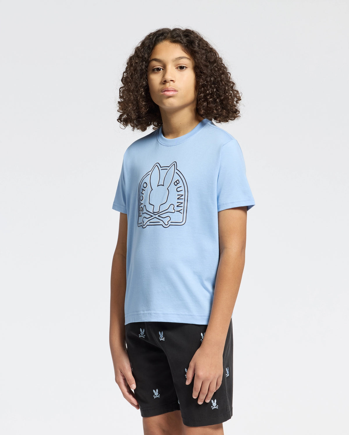 A curly-haired person stands against a plain background wearing Psycho Bunny's eco-friendly KIDS MELBOURNE GRAPHIC TEE in light blue, made from Peruvian Pima cotton, featuring a bunny and crossbones design. They also wear black shorts with a small repeating pattern.